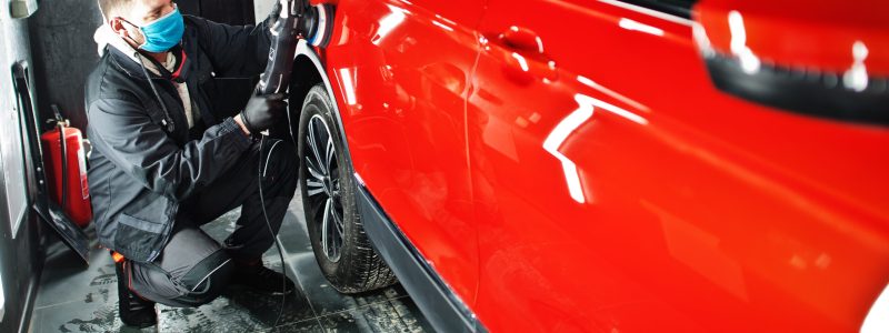 Car detailing concept. Man in face mask with orbital polisher in repair shop polishing orange suv car.